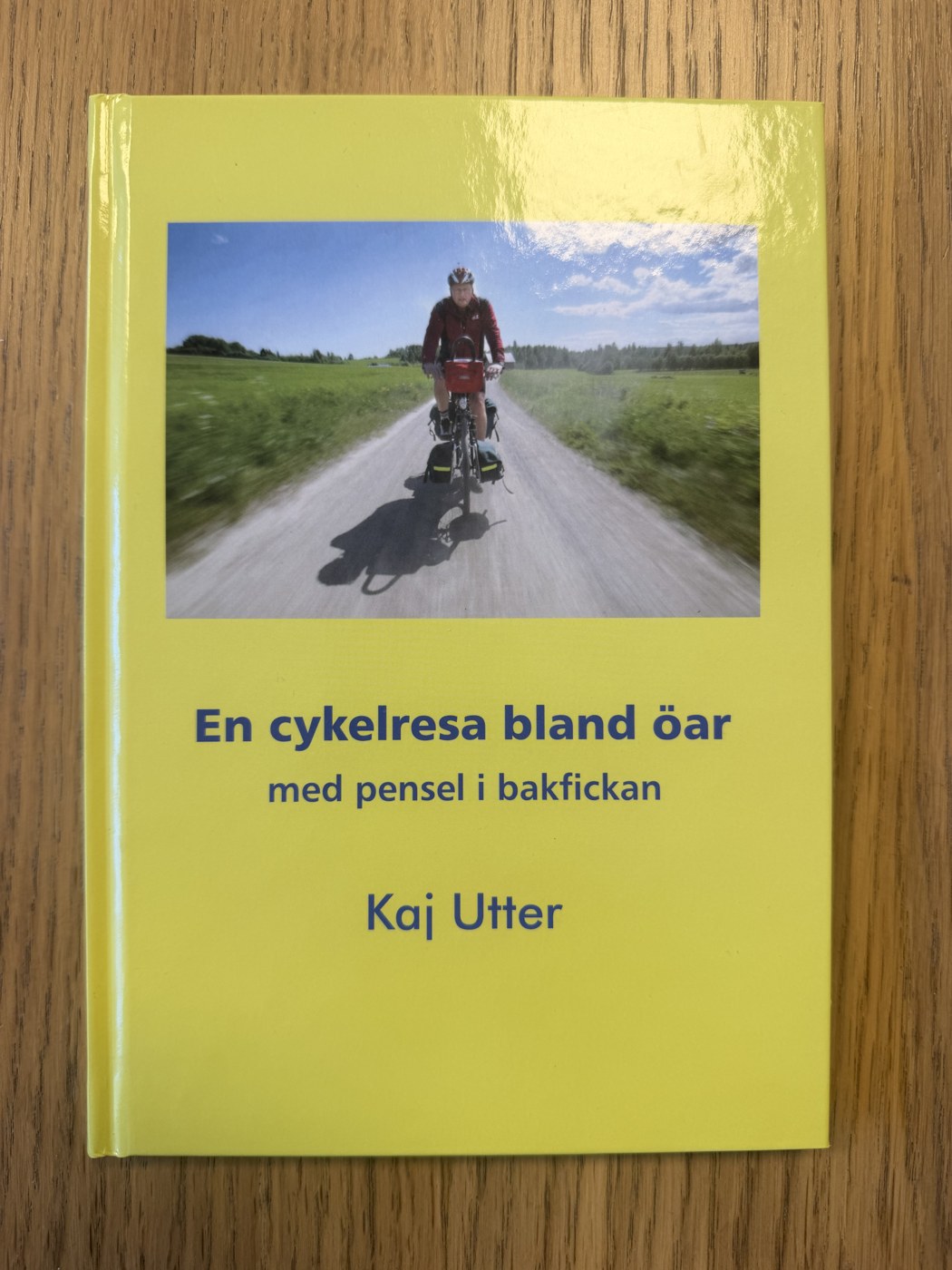 Gult bokomslag med en cyklist på en grusväg mellan gröna ängar